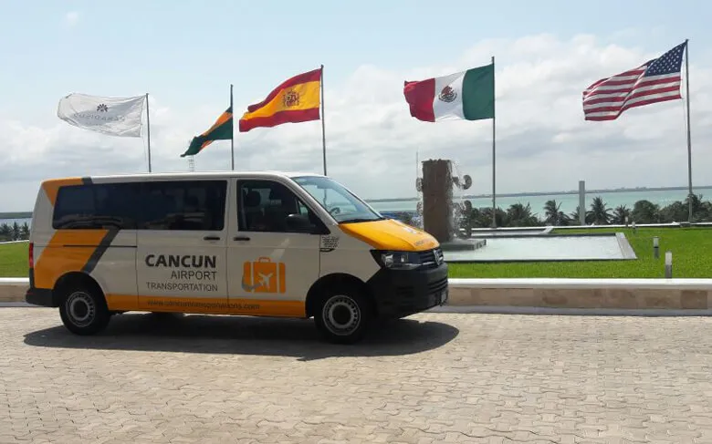 Van labeled with Transportasi Bandara Cancun corporate image parked in front of a water fountain located at Paradisus Resort with several flags, including United States, Spain and Mexico flags. A lagoon, blue sky and some clouds on the background.